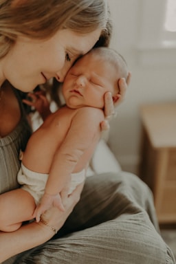 A tender moment between a mother and her newborn baby, with gentle expressions of love and closeness. The baby is peacefully asleep in the mother's arms, who is holding the infant gently against her chest. Soft lighting adds to the intimate ambiance.