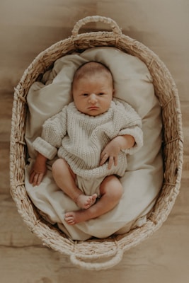 A newborn baby is resting in a woven basket, clad in a knitted cream-colored outfit. The basket is lined with soft fabrics, providing a cozy setting. The baby's expression is calm and serene, positioned comfortably with tiny hands and feet visible.