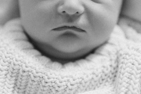 A close-up, black and white photograph of a baby's face, focusing on the lips and part of the nose. The baby is wearing a textured knit sweater, which adds a soft visual element to the image.