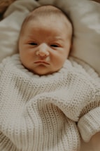 A close-up of a newborn baby wearing a soft, knitted sweater, lying on a light-colored blanket. The baby appears calm and relaxed.