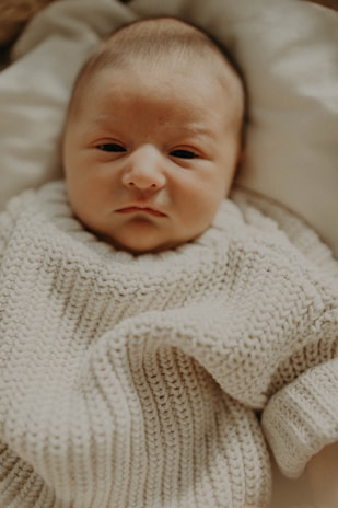 A close-up of a newborn baby wearing a soft, knitted sweater, lying on a light-colored blanket. The baby appears calm and relaxed.