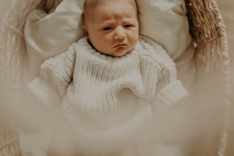 A baby dressed in a cozy, cream-colored knitted sweater lies in a woven basket. The light is soft and warm, creating a calm and gentle atmosphere.