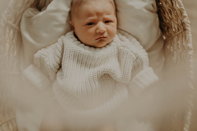 A baby dressed in a cozy, cream-colored knitted sweater lies in a woven basket. The light is soft and warm, creating a calm and gentle atmosphere.