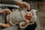 A newborn baby dressed in a knitted beige outfit and a large white headband is cradled in an adult's arms. The baby's eyes are closed, suggesting a peaceful or sleeping state, while the adult's hands gently support the baby.