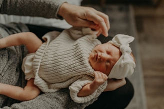 A newborn baby dressed in a knitted beige outfit and a large white headband is cradled in an adult's arms. The baby's eyes are closed, suggesting a peaceful or sleeping state, while the adult's hands gently support the baby.
