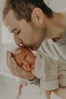 A close-up of a man gently kissing the head of a newborn baby who is wrapped in a knitted sweater. The man holds the baby tenderly in his hands, expressing a strong sense of warmth and affection.