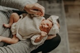 A newborn baby wearing a cozy knitted outfit and a large white bow headband is cradled in someone's arms. The baby's eyes are closed, and they appear to be sleeping peacefully, creating a serene and tender mood.