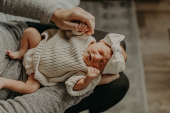 A newborn baby wearing a cozy knitted outfit and a large white bow headband is cradled in someone's arms. The baby's eyes are closed, and they appear to be sleeping peacefully, creating a serene and tender mood.