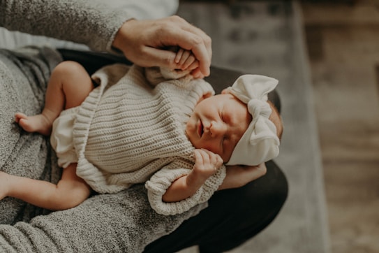 A newborn baby wearing a cozy knitted outfit and a large white bow headband is cradled in someone's arms. The baby's eyes are closed, and they appear to be sleeping peacefully, creating a serene and tender mood.