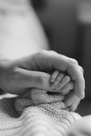 Softly lit close-up of a mother tenderly holding her baby's hand, surrounded by green leaves.