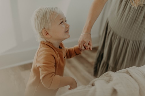 A young child with blonde hair, wearing a brown sweater, is looking up with a joyful expression while holding hands with an adult. The background is softly blurred, featuring natural light and a neutral-colored setting.