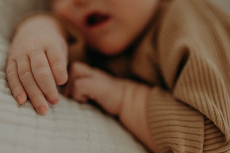 A close-up of a baby's hand resting gently on a soft, textured surface. The baby's outfit is made of a ribbed, earthy-toned fabric, contributing to a cozy and serene atmosphere.