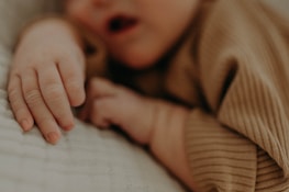 A close-up of a baby's hand resting gently on a soft, textured surface. The baby's outfit is made of a ribbed, earthy-toned fabric, contributing to a cozy and serene atmosphere.