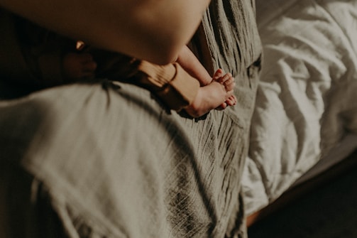 A close-up view of a tiny baby's feet held by an adult's hand, resting against a soft, textured fabric. The background includes a bed with white sheets, providing a warm and gentle ambiance.