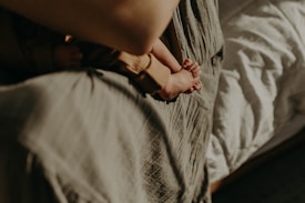 A close-up view of a tiny baby's feet held by an adult's hand, resting against a soft, textured fabric. The background includes a bed with white sheets, providing a warm and gentle ambiance.
