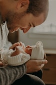 A tender moment is captured with a man lovingly holding a sleeping newborn baby. The baby is dressed in a cozy knitted outfit and a soft, white hat. The man, with a gentle expression, closely observes the baby, creating an intimate and serene atmosphere.