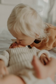 A young child gently kisses a baby on the forehead while laying on a bed. Both have blonde hair and are wearing soft, warm clothing. The scene is cozy and intimate, with a blurred background that adds to the softness of the moment.