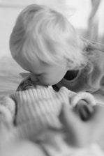 A child with light hair gently kisses a newborn baby lying down, wrapped in a textured blanket. The black and white image captures a tender and intimate moment between siblings in a serene setting.
