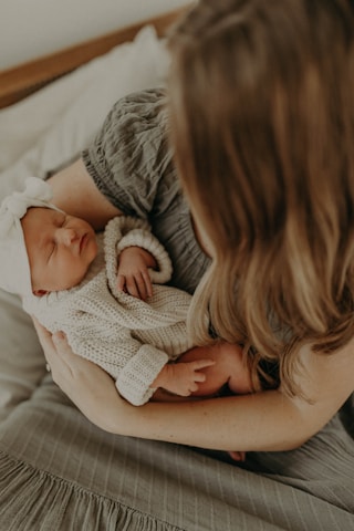 A woman is gently cradling a sleeping baby in her arms. The baby is dressed in a soft knitted outfit and wears a white headband with a bow. The setting is warm and intimate, suggesting a peaceful and nurturing environment.