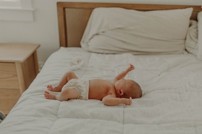 A baby in a diaper lies on a large, white quilted bed with light wood furniture in the background. The baby appears relaxed, with arms stretched and head turned to the side.