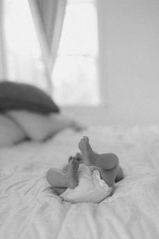 A black and white photo featuring a baby lying on its back on a bed. The baby's legs and feet are playfully lifted in the air. The scene appears soft and serene, with light streaming in from a blurred background.
