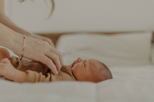 A peaceful baby lies on a soft surface wearing a brown outfit. A hand gently touches the baby's chest, conveying a sense of care and tenderness.
