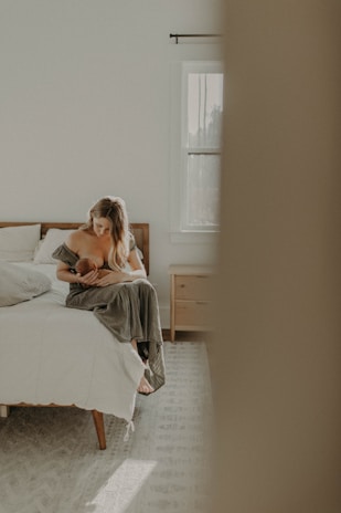A serene, softly lit bedroom features a woman sitting on the edge of a bed while cradling a baby. The room has a cozy and minimalistic design with a light color palette, including a window letting in natural light, light-colored walls, and wooden furniture.