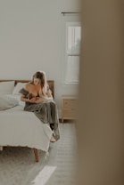 A serene, softly lit bedroom features a woman sitting on the edge of a bed while cradling a baby. The room has a cozy and minimalistic design with a light color palette, including a window letting in natural light, light-colored walls, and wooden furniture.