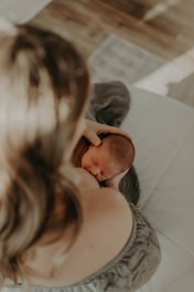 A newborn baby is gently cradled against an adult's chest, with the adult's hands supporting the baby's head. The setting appears soft and serene, with natural light casting gentle shadows.