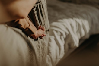 A baby's feet resting on a soft surface, with a blurred warm-toned background. The setting suggests warmth and comfort, possibly a home environment.