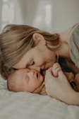 A tender moment between a mother and her sleeping baby, as they gently touch foreheads while the mother holds the baby's hand. The mood is serene and intimate, with soft lighting enhancing the sense of warmth and closeness.