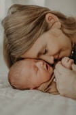 A tender moment between a woman and a sleeping baby as they rest their heads together, conveying love and warmth. The woman has her eyes closed, smiling gently, while the baby sleeps peacefully, wearing a soft, brown outfit. Their hands are tenderly clasped together, enhancing the intimate atmosphere.