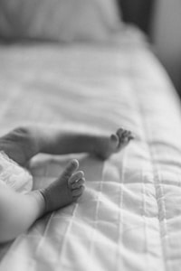 A black and white photograph of a baby lying on a soft, quilted surface. The focus is on the small feet and toes of the infant, which adds a delicate and tender quality to the image.