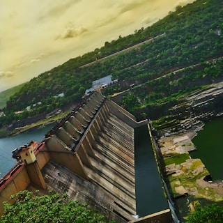 A large dam structure stands amidst a lush, green landscape with water flowing beneath it. The dam is constructed from concrete and features a series of vertical gates. The surrounding area includes hills covered with dense vegetation and a partly cloudy sky with a warm hue.