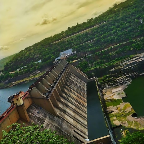 A large dam structure stands amidst a lush, green landscape with water flowing beneath it. The dam is constructed from concrete and features a series of vertical gates. The surrounding area includes hills covered with dense vegetation and a partly cloudy sky with a warm hue.