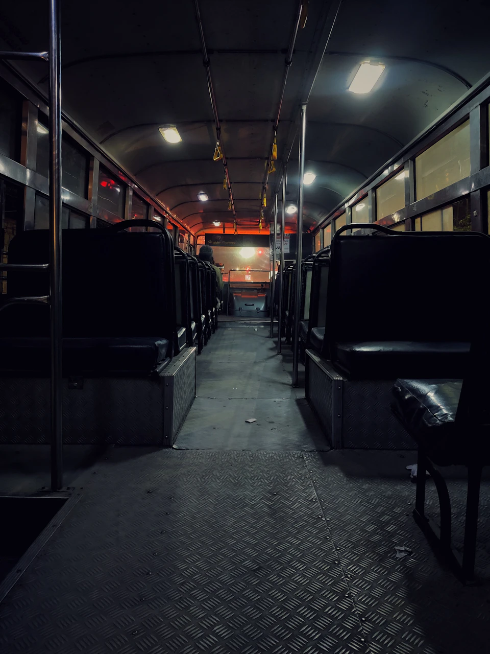 The image depicts a dimly lit bus interior, with rows of black seats facing each other and a single aisle running down the center. The floor is covered in a shiny metal grate that reflects the soft glow from the ceiling lights. A solitary figure can be seen standing near the front of the bus, adding an element of mystery to the scene.
