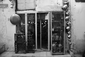 A black-and-white image of a small shop entrance with large glass doors and a glass display case. Various metal kitchenware items, such as kettles and pots, are hanging on the wall. Inside the glass case, more metal utensils and a traditional samovar are displayed. The rough texture of the shop's exterior wall and the assortment of metal gas burners stacked outside add to the rustic ambiance.
