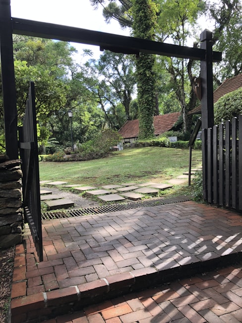 A garden landscape with a stone path leading through a green lawn, surrounded by lush trees and vegetation. The scene is viewed from beneath a dark wooden archway with a brick patio in the foreground and a charming building with a sloped roof in the background.