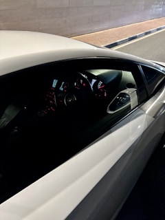 A close-up view of the interior of a car from the passenger side, highlighting the dashboard and steering wheel with illuminated dials. The exterior view shows a beige or light-colored road wall and sidewalk alongside the car.