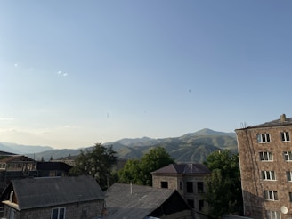 A residential area with several buildings in the foreground, surrounded by green trees. In the background, there are rolling hills with gentle slopes under a clear blue sky. Birds are flying overhead, adding movement to the scene.