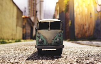 A toy model of a vintage van is placed on a paved road, with blurred urban buildings and walls in the background. The perspective is low to the ground, making the van appear larger than it is. The scene is illuminated by warm sunlight, casting a soft glow.