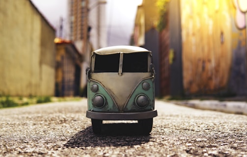 A toy model of a vintage van is placed on a paved road, with blurred urban buildings and walls in the background. The perspective is low to the ground, making the van appear larger than it is. The scene is illuminated by warm sunlight, casting a soft glow.