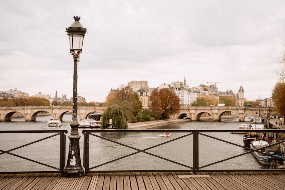 The view from the wooden deck and railings of the Pont des Arts towards the stone arches of the Pont Neuf and the tip of the Île de la Cité, under a flat grey sky, with an old Paris street lamp in the foreground