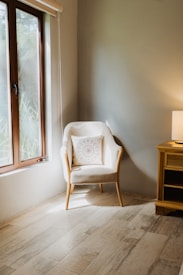 A cozy, well-lit corner of a room features a comfortable armchair with a decorative pillow. Next to the chair is a wooden side table with a lamp casting a warm glow. Light streams through a window, accentuating the natural tones of the wooden floor.