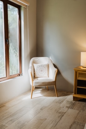 A cozy, well-lit corner of a room features a comfortable armchair with a decorative pillow. Next to the chair is a wooden side table with a lamp casting a warm glow. Light streams through a window, accentuating the natural tones of the wooden floor.