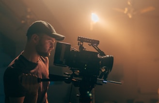 A person is operating a professional video camera on a set, surrounded by warm, soft lighting and a slightly misty atmosphere. The individual is wearing a hat and focused intently on the camera.