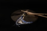 A pair of drumsticks rest on a bronze cymbal, accompanied by a tassel with blue and white threads. The background is dark, emphasizing the metallic sheen of the cymbal and the wood texture of the drumsticks.