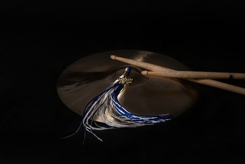 A pair of drumsticks rest on a bronze cymbal, accompanied by a tassel with blue and white threads. The background is dark, emphasizing the metallic sheen of the cymbal and the wood texture of the drumsticks.