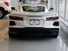 A sleek white sports car is parked indoors, showcasing an aerodynamic design with sharp angles and stylish rear lights. The car features a license plate from a dealership named White Allen in Dayton, Ohio. It is positioned on a shiny tiled floor inside a car showroom, with another vehicle partially visible on the left side.