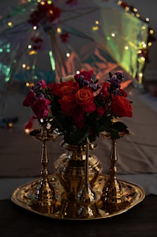 A richly decorated floral arrangement features vibrant red and pink flowers placed in a golden vase, accented with intricate metallic candle holders. Behind the flowers, a multi-colored, illuminated umbrella adds a festive and magical atmosphere with its glowing lights.
